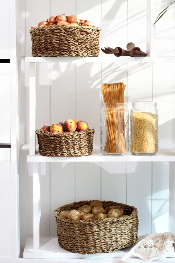organized-pantry-shelf-with-fresh-produce-31390646 A neatly organized pantry shelf with wicker baskets holding apples, peaches, and potatoes, alongside pasta and grains.